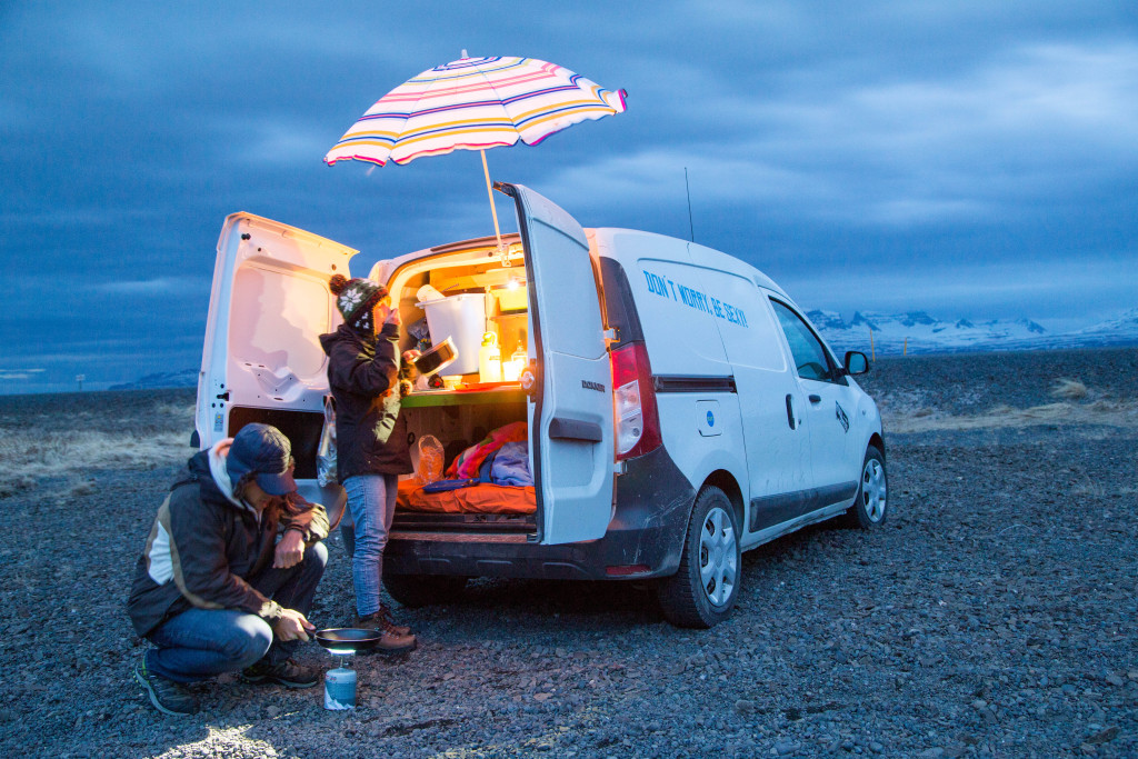 KuKu Campervan Cooking Setup on the Icelandic Ring Road A couple cooking beside their KuKu campervan under a striped umbrella during an Iceland road trip – perfect example of van life freedom and creativity in unpredictable weather.
