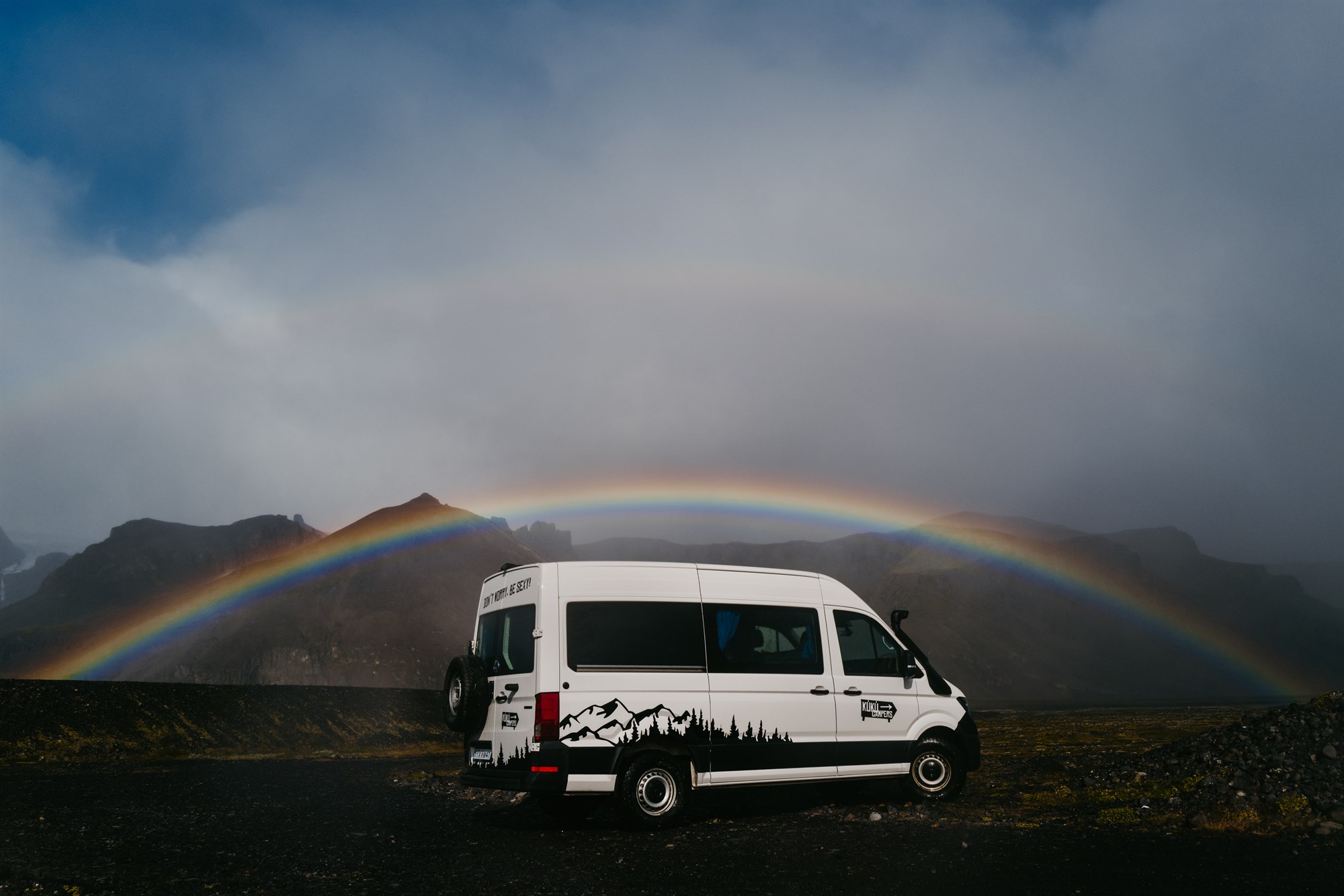 A scenic view of a campervan parked in the wild Icelandic landscape, with a stunning rainbow arching over the mountains. The perfect road trip moment!